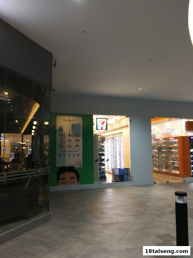The storefront of a 7-Eleven convenience store, featuring its logo and a promotional display, within a shopping mall.