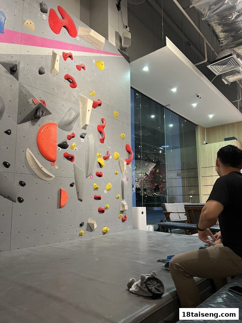 An indoor bouldering gym featuring a climbing wall with colorful holds and a person resting nearby.