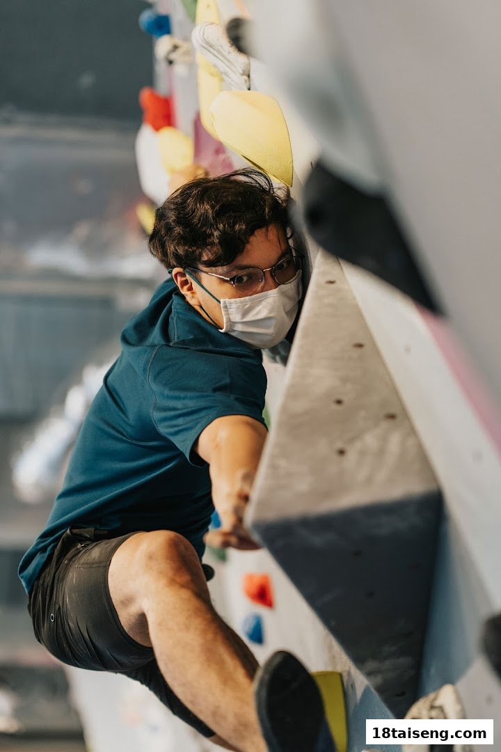 A person wearing a mask and glasses is actively bouldering on a climbing wall with colorful holds.