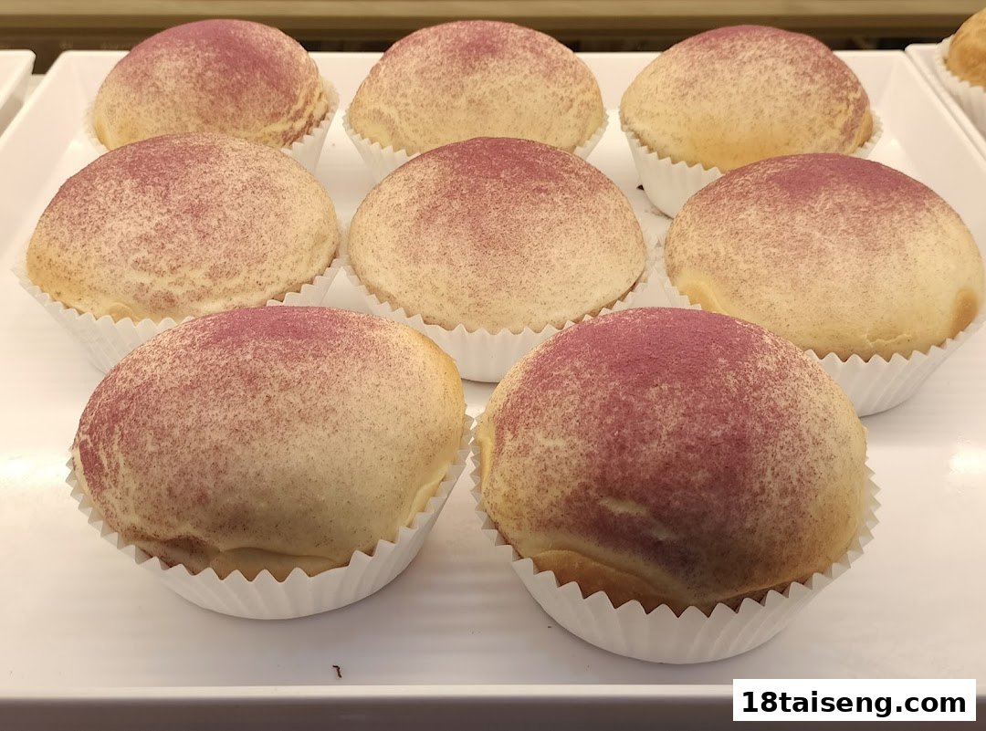 A tray of freshly baked, round buns or pastries, lightly dusted with purple powder, displayed in white paper cups.