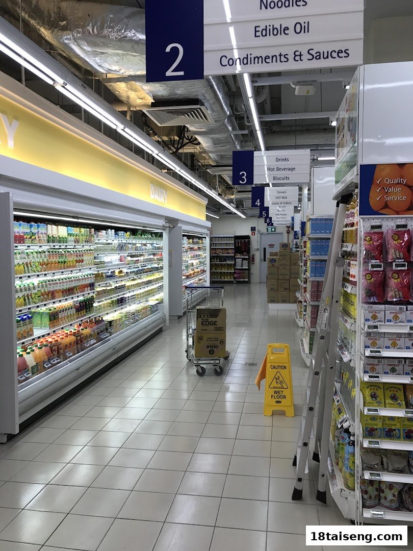 A brightly lit supermarket aisle at FairPrice Tai Seng, featuring refrigerated drinks, dairy, and various grocery sections.