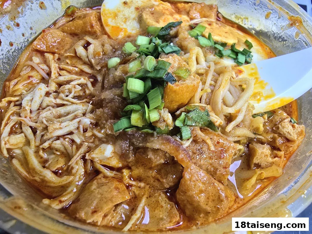 A close-up of a vibrant bowl of curry noodles or laksa, topped with shredded chicken, tofu puffs, and spring onions.