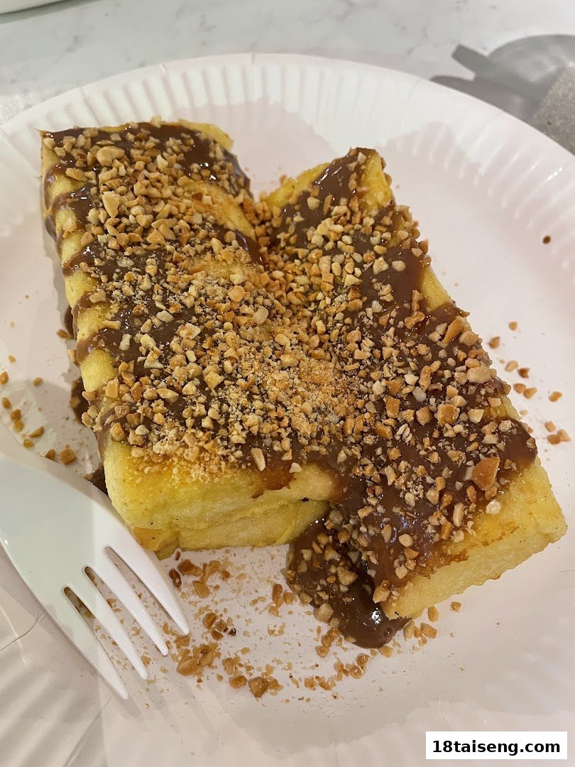 Two pieces of golden toast generously topped with chocolate sauce and crushed peanuts on a white paper plate.