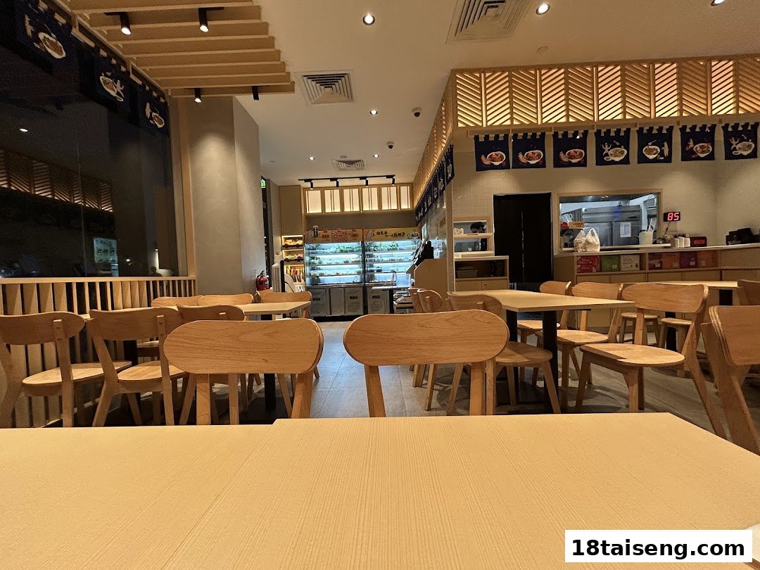 Interior view of a modern, brightly lit restaurant with wooden tables and chairs, and a visible kitchen area.