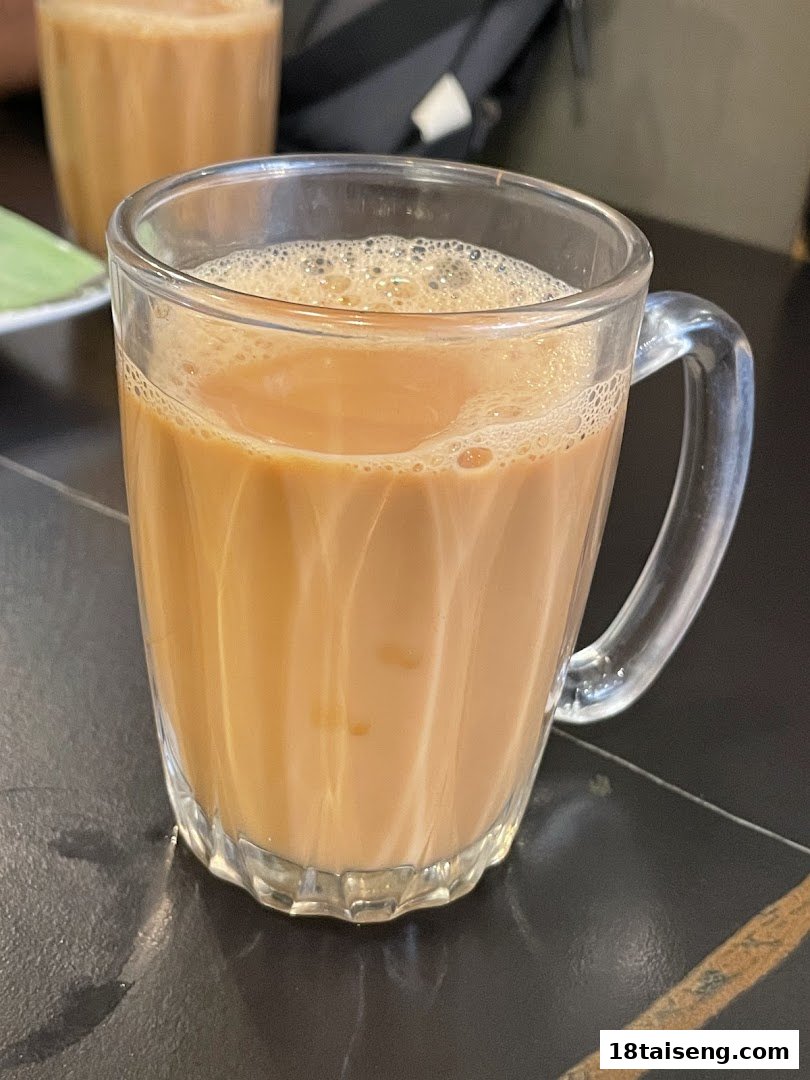 A close-up of a frothy glass of milk tea or coffee on a dark table at Jalan Kayu The Prata Cafe.