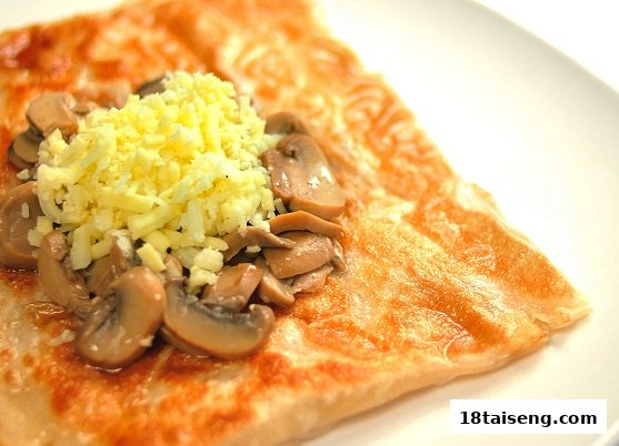 A golden-brown prata topped with sautéed mushrooms and shredded cheese on a white plate.