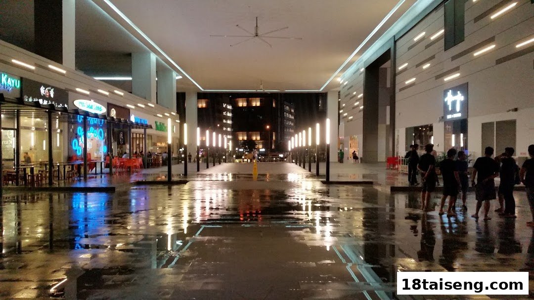 Illuminated shopfronts and a wet plaza at night, featuring Jalan Kayu The Prata Cafe.