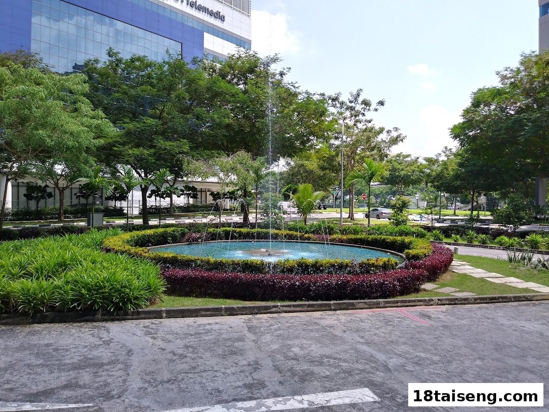 A vibrant outdoor scene featuring a circular fountain surrounded by lush greenery and a modern building.