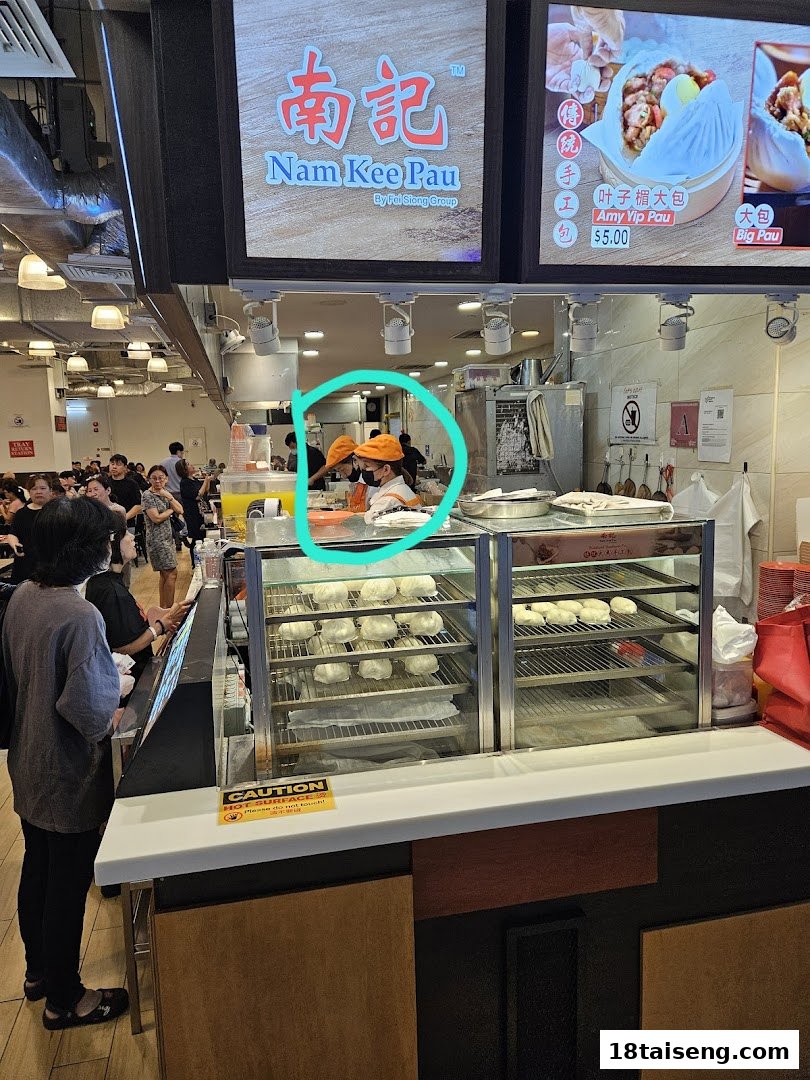 A bustling food stall counter at Nam Kee Pau, featuring steamed buns in display cases and staff serving customers.