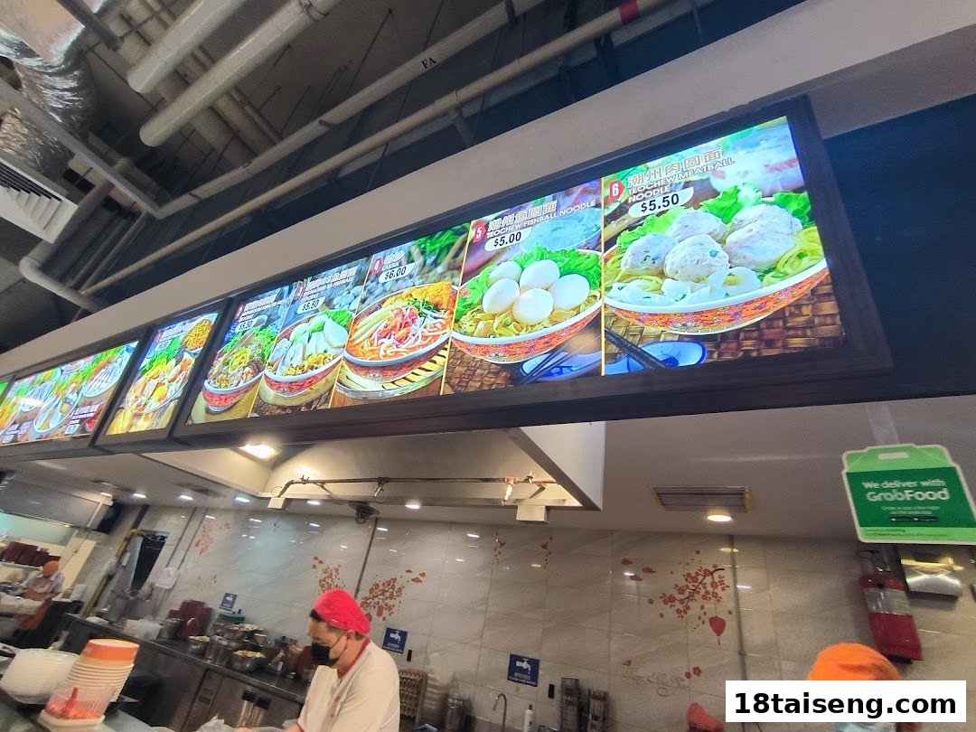 Illuminated menu boards displaying various noodle dishes and prices at a food stall.