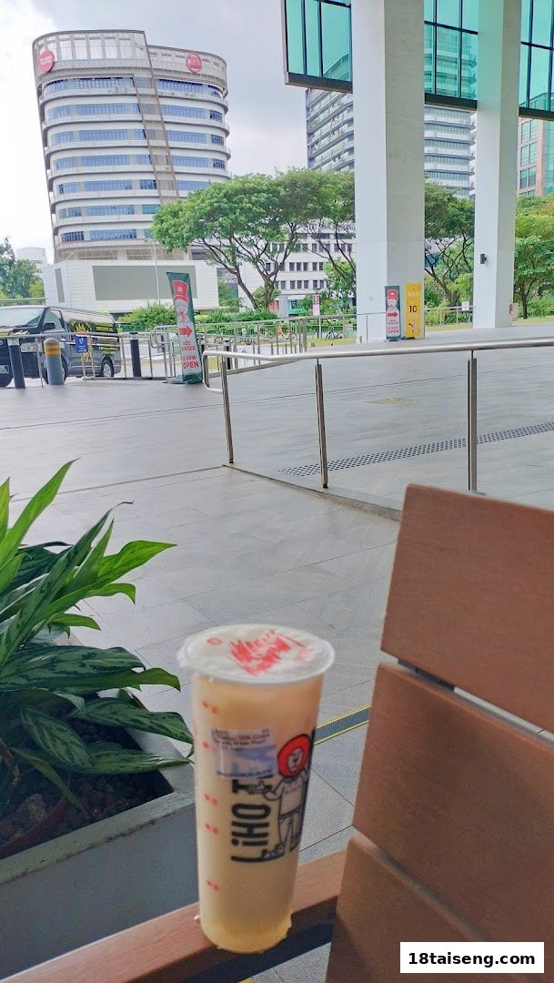 A LiHO TEA bubble tea drink sits on a bench with a modern building and outdoor area in the background.