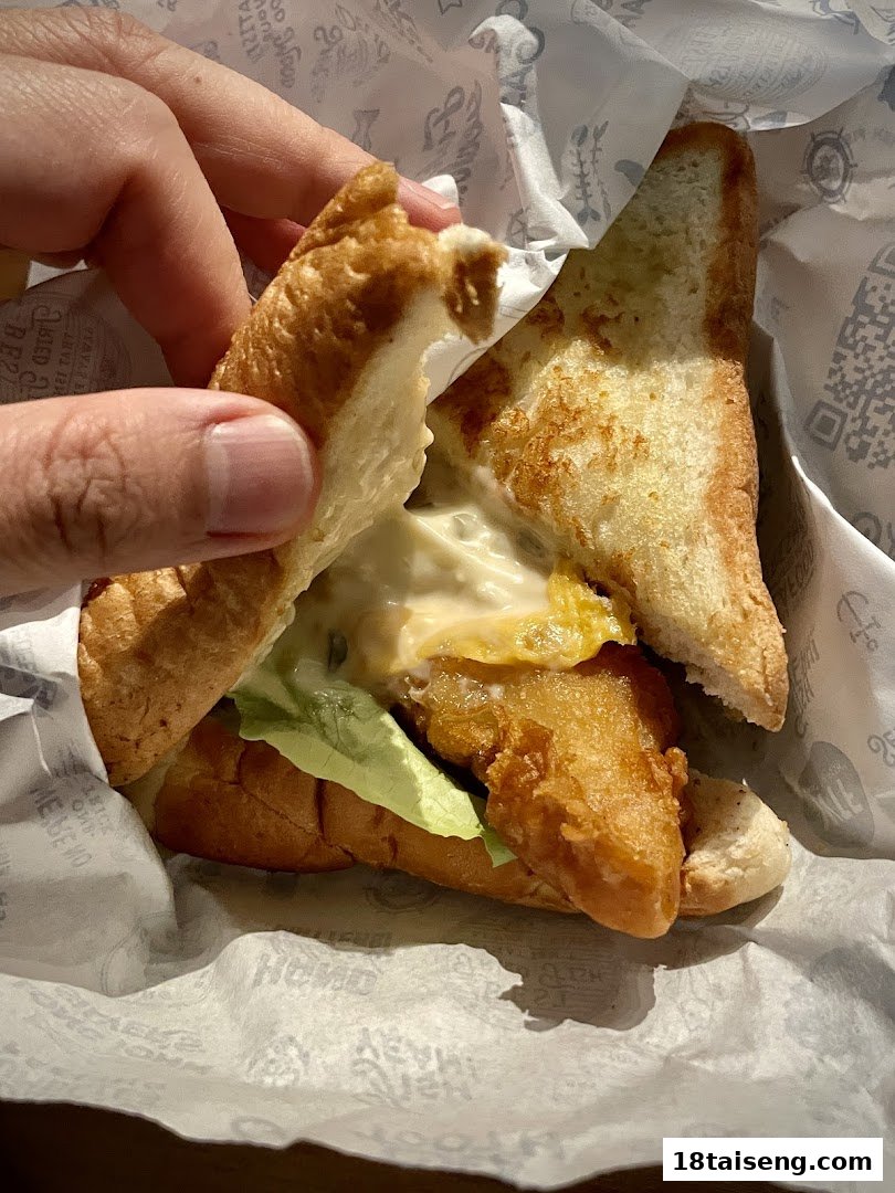 A hand holds a delicious fish sandwich with lettuce and tartar sauce, wrapped in branded paper.