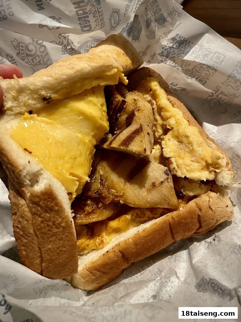 A close-up of a hearty sandwich filled with grilled chicken and scrambled eggs, wrapped in branded paper.