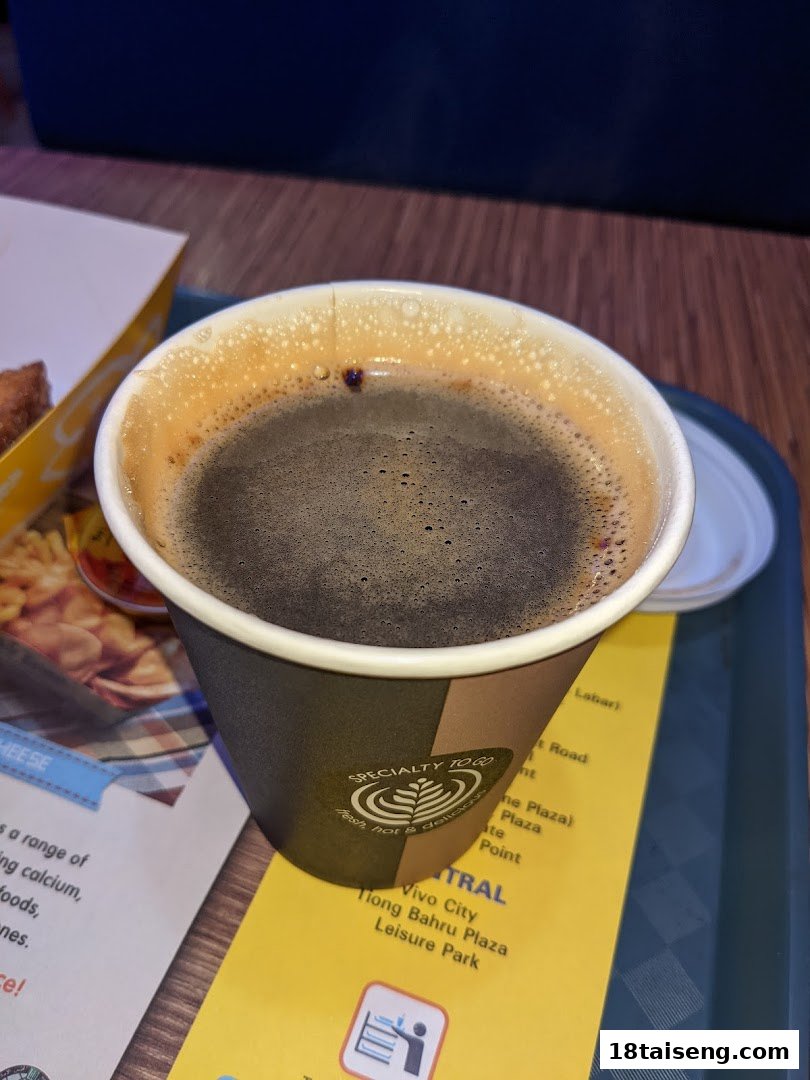 A close-up of a hot coffee cup with food in the background on a tray.