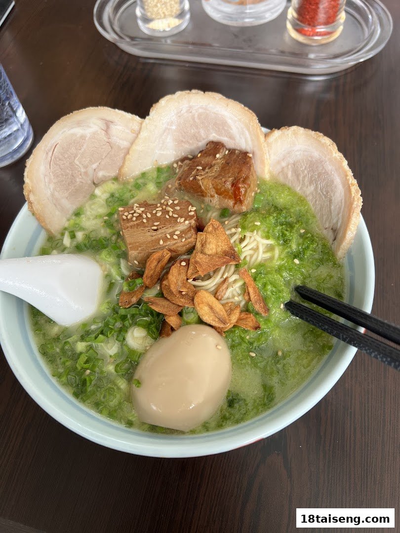 A close-up of a delicious ramen bowl featuring green broth, pork slices, braised pork, egg, and crispy garlic.