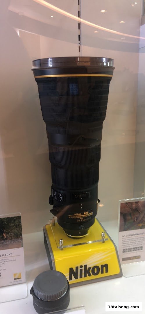 A large Nikon camera lens displayed on a yellow stand in a retail store.