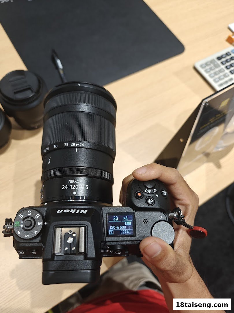 A hand holds a Nikon Z series camera with a NIKKOR 24-120mm f/4 S lens, showing its display.