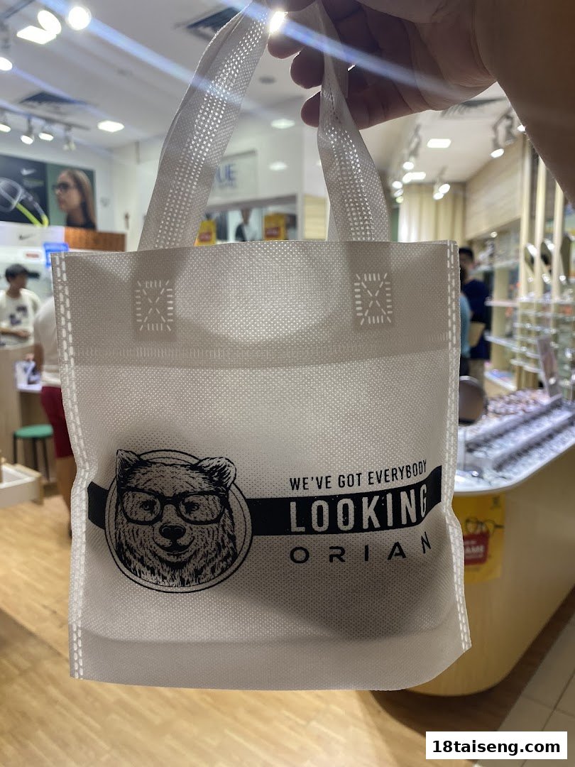 Hand holding a white Orian shopping bag with a bear logo inside an optical store.