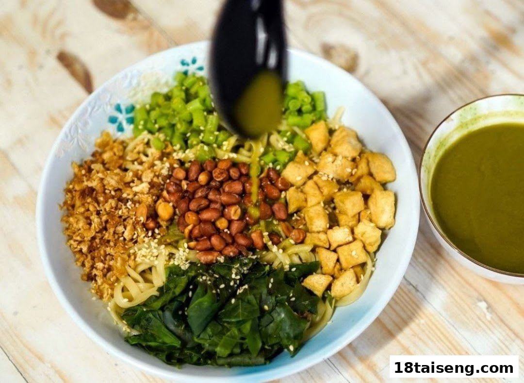 A vibrant bowl of vegetarian noodles with peanuts, tofu, greens, and a green sauce being poured.