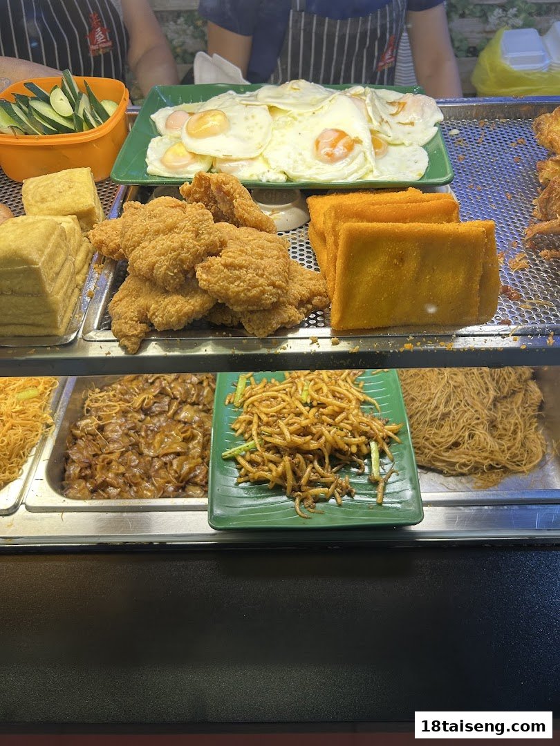 A display of various cooked dishes including fried chicken, eggs, and noodles at a food stall.