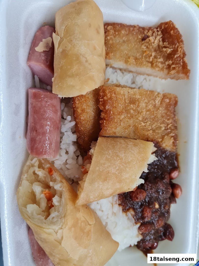A takeout meal featuring rice, fried items, sausages, and a spring roll in a white container.