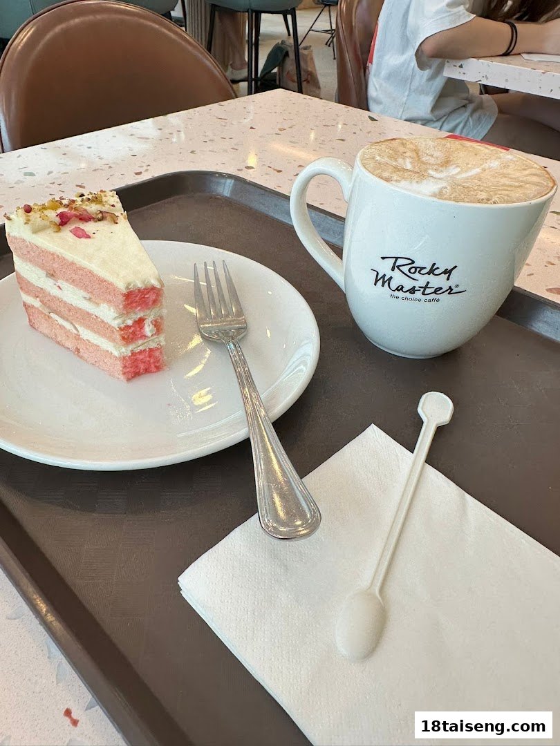 A slice of pink layered cake with a fork and a Rocky Master coffee mug on a tray.