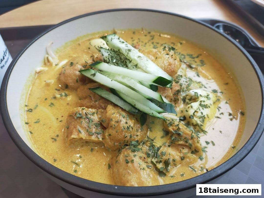 A vibrant bowl of curry noodles, possibly laksa, topped with tofu puffs, cucumber, and herbs.