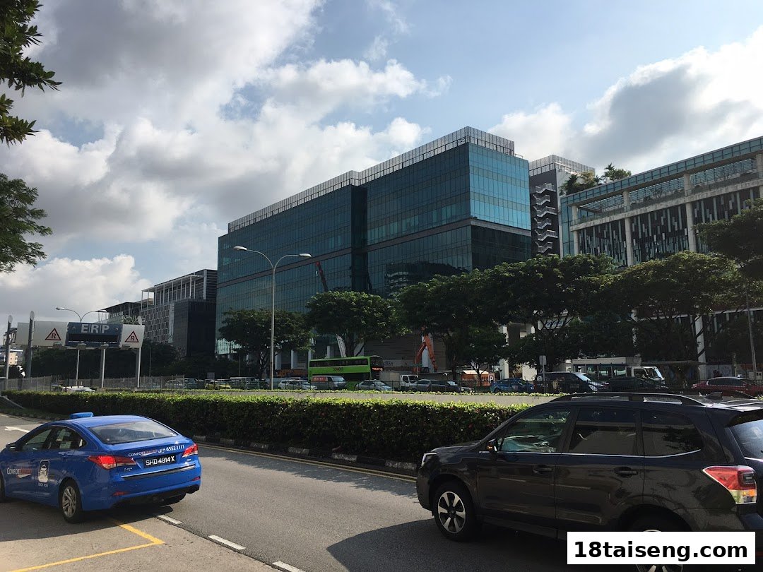 A modern office building complex with reflective glass facades, cars, and lush trees under a cloudy sky.