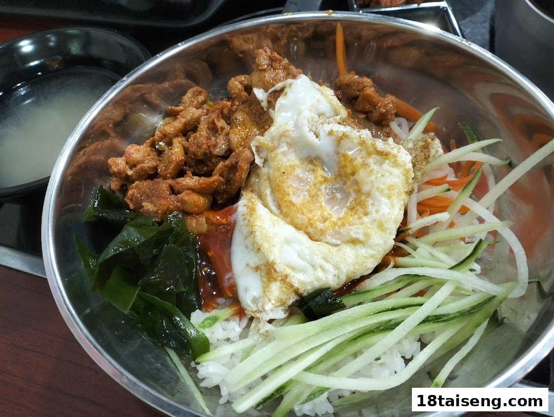 A vibrant Korean rice bowl with spicy pork, fried egg, fresh vegetables, and rice in a metal bowl.