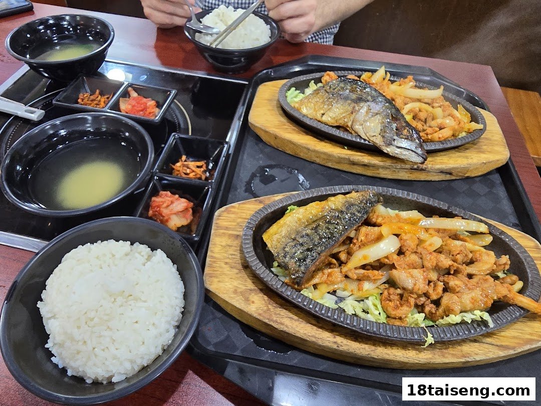 A table set with two sizzling Korean dishes featuring fish, spicy pork, rice, soup, and banchan.