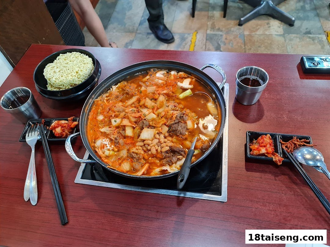 A large pot of bubbling Korean stew with noodles and banchan on a wooden table.