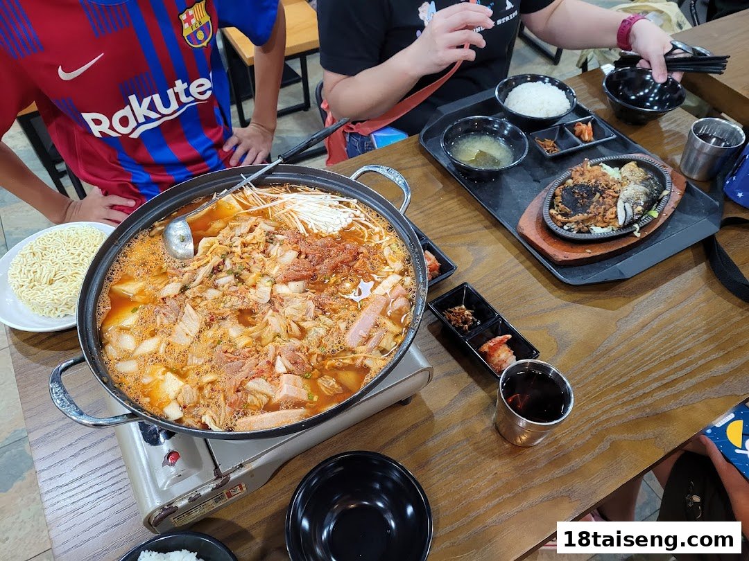 A large pot of bubbling Korean kimchi stew and other dishes on a wooden table, with diners.