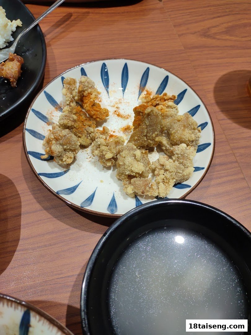 A plate of crispy fried chicken with seasoning, alongside a bowl of soup, on a wooden table.