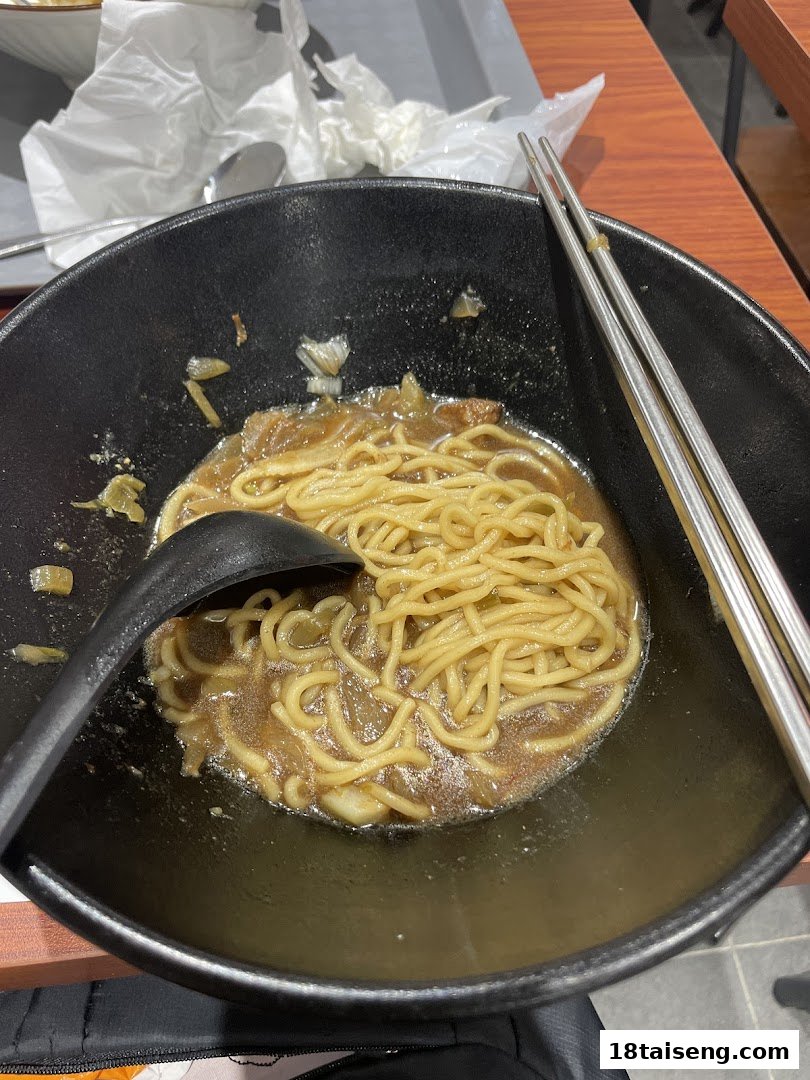A close-up of a bowl of savory noodles with broth, a spoon, and chopsticks on a table.