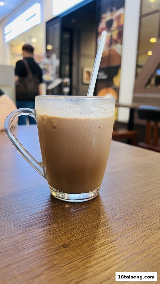 A clear glass mug filled with a brown beverage and a straw sits on a wooden table.