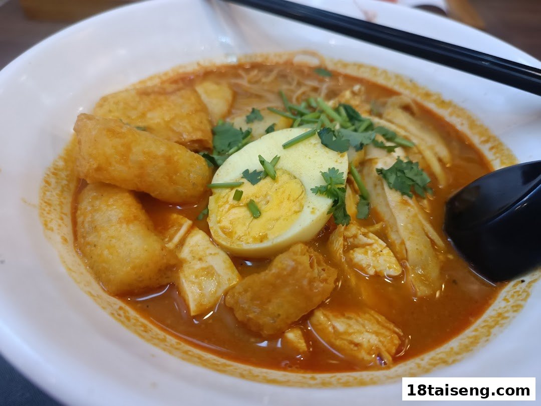 A close-up of a vibrant bowl of curry noodles or laksa, garnished with egg, chicken, and fried tofu puffs.