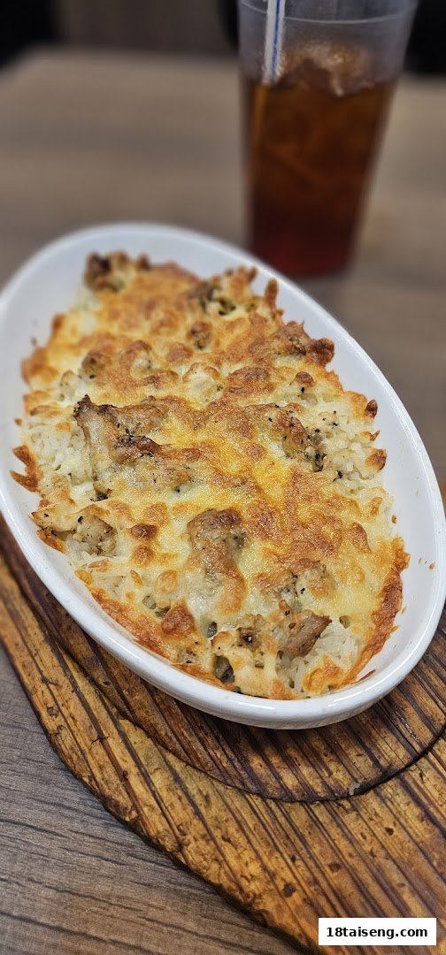 A close-up of a baked cheesy rice dish with meat, served on a wooden board, alongside an iced tea.