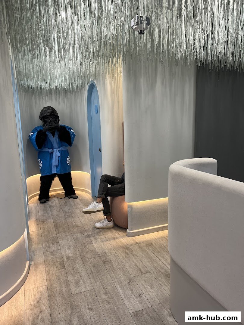 A person in a gorilla costume stands in a modern waiting area with a person seated.