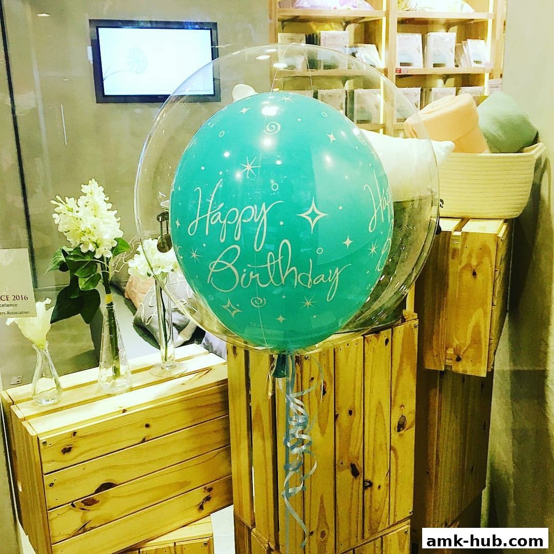 A teal 'Happy Birthday' balloon in a clear bubble, displayed with flowers and wooden crates.
