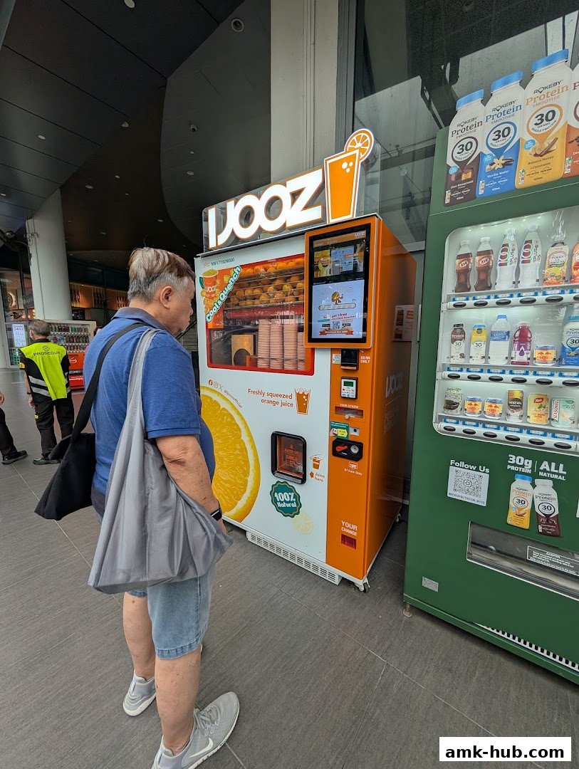 A man stands in front of an orange juice vending machine and a green beverage vending machine.
