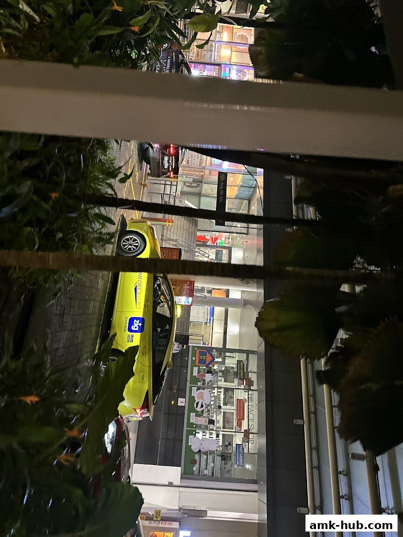 A yellow taxi is parked outside a brightly lit shop with various products displayed in the window.