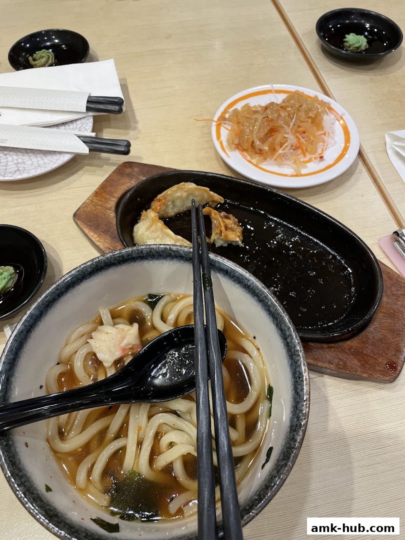 A close-up of a table at Ichiban Sushi Ang Mo Kio with udon, gyoza, and side dishes.