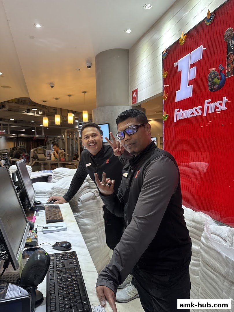 Two smiling staff members at the Fitness First reception desk, one making a peace sign.