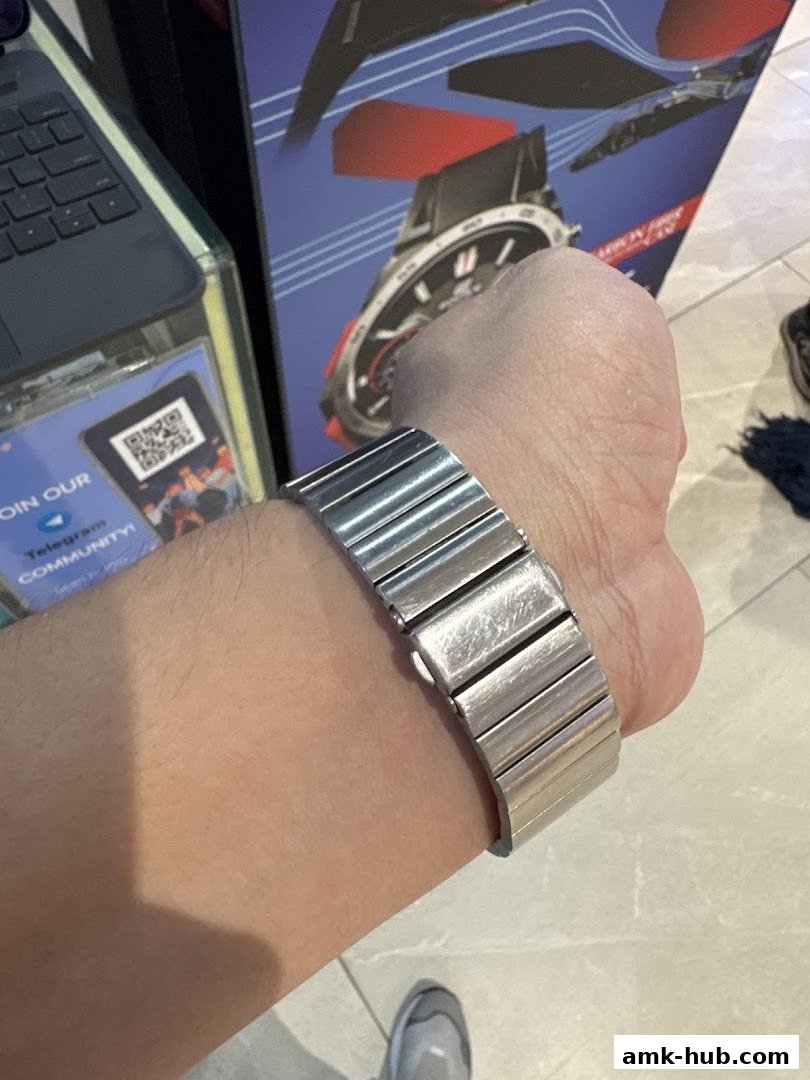 A person's wrist wearing a metal watch band with a watch advertisement in the background.