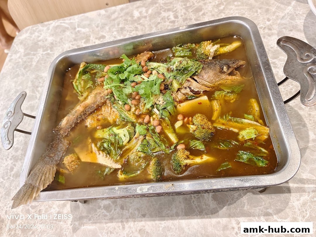 A whole fish dish with vegetables and peanuts in a spicy broth served in a metal tray.