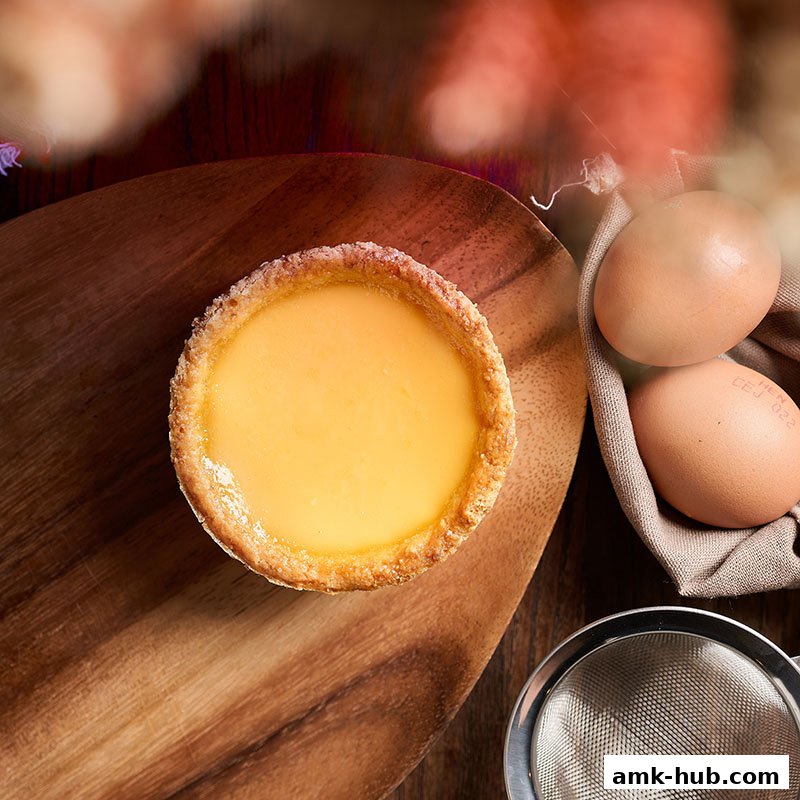 A close-up of a creamy egg tart on a wooden board with fresh eggs and a sieve.