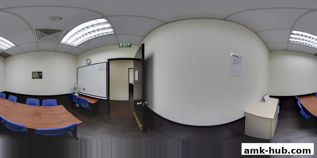 A classroom or meeting room with a large table, chairs, whiteboard, and exit sign.
