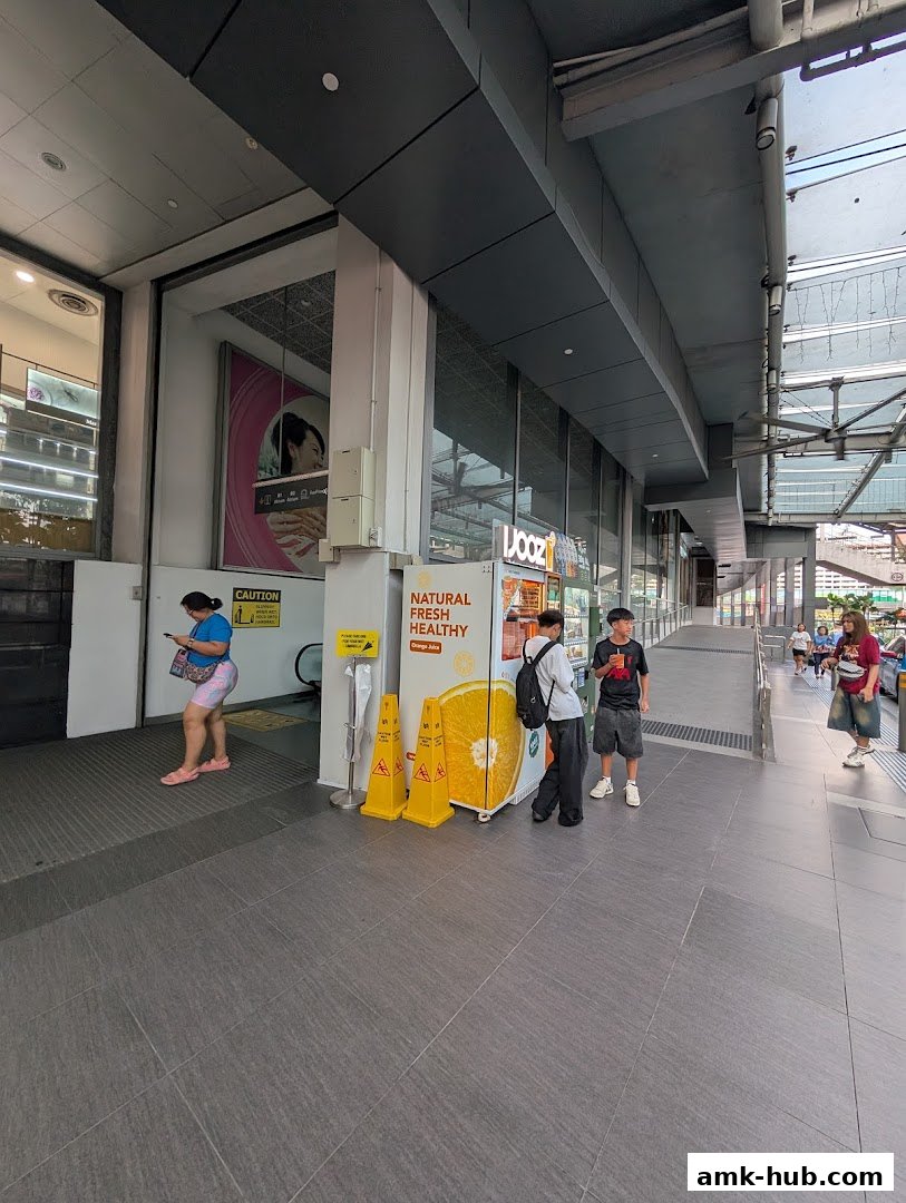 People walking past a juice shop with a large orange advertisement and a vending machine.