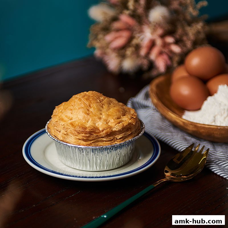A golden-brown chicken pie sits on a plate with a fork and ingredients for baking.