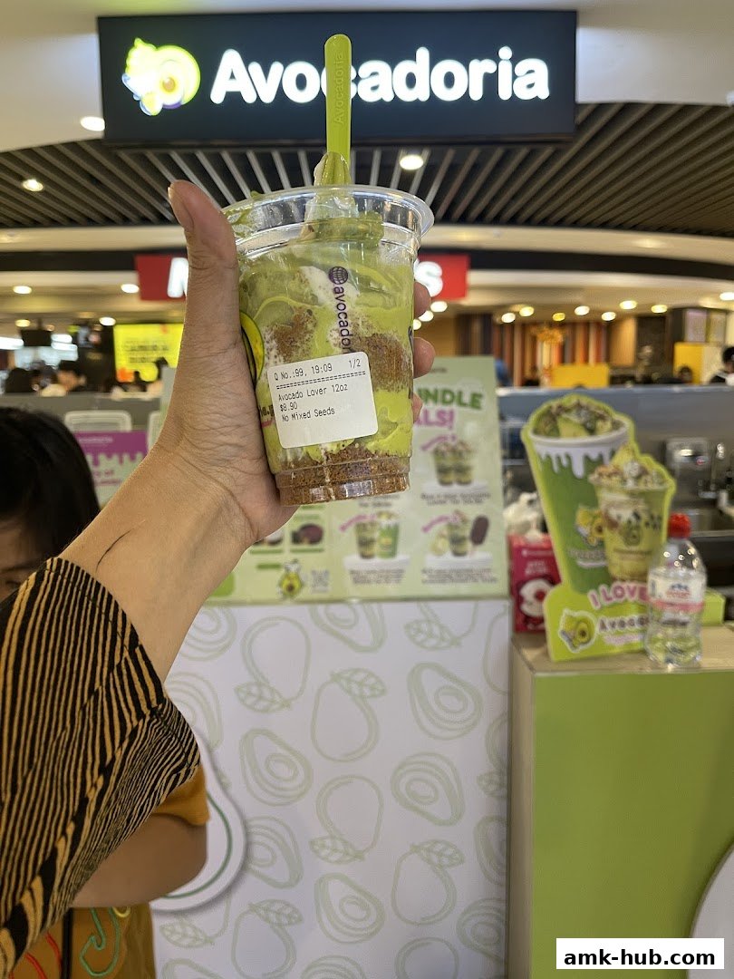 A hand holds a layered avocado dessert in front of the Avocadoria shop sign.